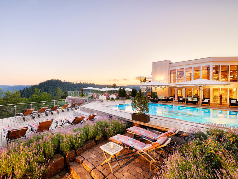 Ein Außenpool mit Poolterrasse, sowie ein beleuchteter Teil des Wellnesshotels inmitten der grünen Natur des Schwarzwalds.