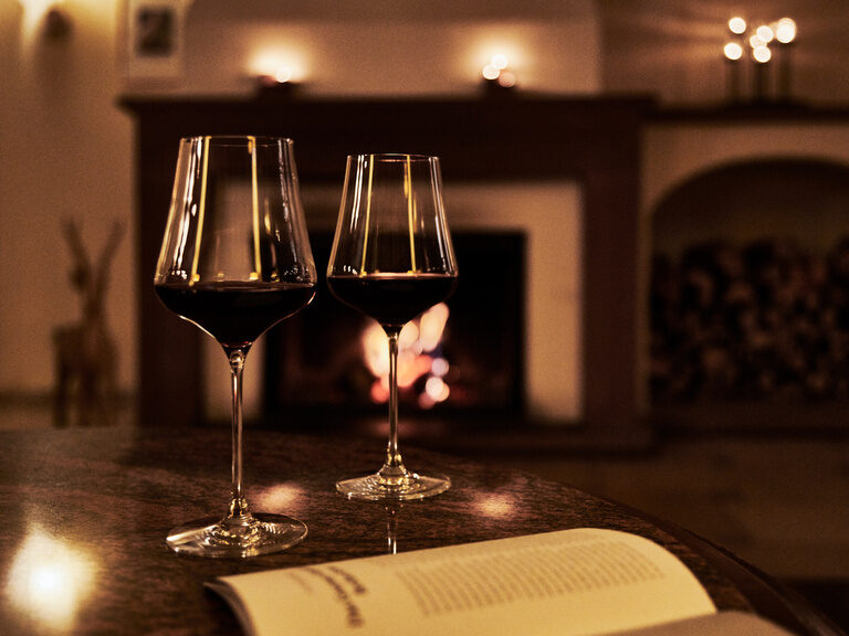Two glasses of red wine stand on a table in front of a lit fireplace.