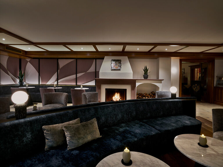 Elegante Hotel-Lounge im Hotel Kronelamm mit Kamin und stilvoller Beleuchtung. Dunkle Farben, gemütliche Sitzgelegenheiten und Marmortische mit Kerzen schaffen eine einladende, luxuriöse Atmosphäre.