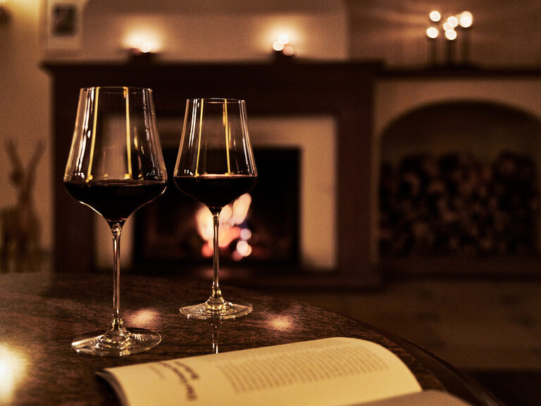 Zwei Gläser Rotwein stehen auf einem Tisch vor einem angefeuerten Kamin