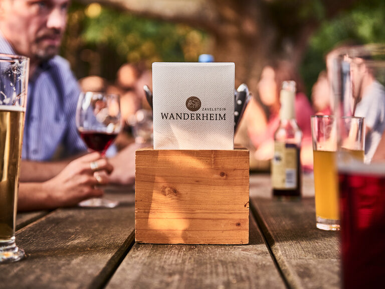 Regionalität spielt im Wanderheim sowohl bei den Speisen als auch bei Getränken eine essentielle Rolle. Nahaufnahme eines Biertisches, auf dem Servietten mit dem Logo des Zavelsteiner Wanderheims und verschiedene Getränke stehen.