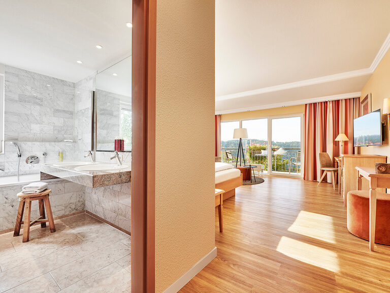 Die Zimmer liegen auf der Südseite im Hotel Kronelamm in den oberen Stockwerken mit Balkon und grandioser Aussicht über den Pool und den Schwarzwald. Ein Blick in das weiße Bad des Deluxe Zimmer Typ Talblick mit Wohnbereich in der rechten Bildhälfte.