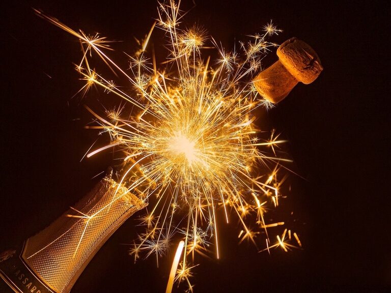 Zwischen dem Hals einer Champagnerflasche und deren Korken erleuchtet ein Feuerwerk.