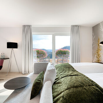 Die Deluxe-Doppelzimmer Talblick liegen sehr ruhig zur Südseite des Hotels Kronelamm in Zavelstein. Ein großzügiges Bett, auf dem weiße Bettwäsche und eine grüne Tagesdecke liegt in einem lichtdurchfluteten Hotelzimmer.