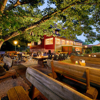 Ein Biergarten mit Tischen und Bänken vor einem roten Haus in Abenddämmerung mit Lichtern.