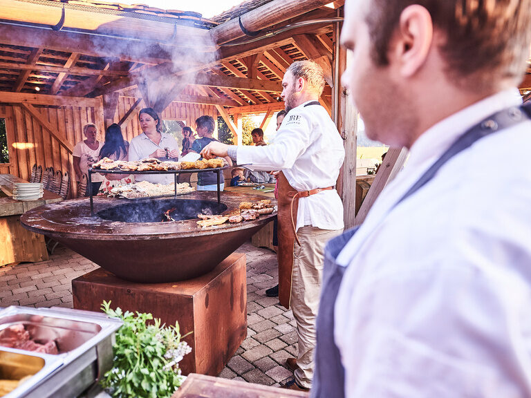 Zwei Köche stehen an einem großen Grill und bereiten, im Rahmen einer Feierlichkeit, leckere Grillspezialitäten zu.