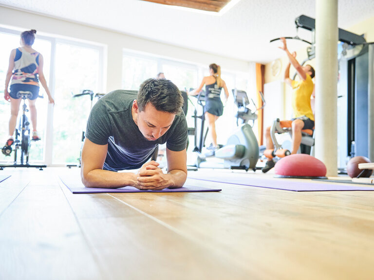 Ein junger Gast trainiert im Ellbogenstütz auf einer Sportmatte in einem Fitnessraum.