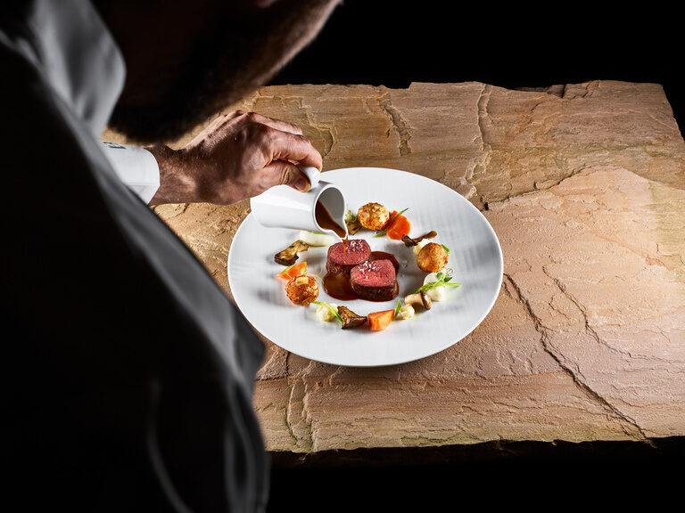 A chef nibbles sauce from a plate of venison and vegetables.