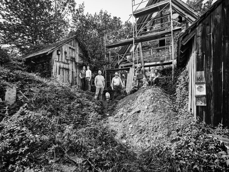 Schwarzweiß Bild mit Gastgeberfamilie Berlin, die zwischen Erdhügeln vor einer Hütte und einem Haus steht.