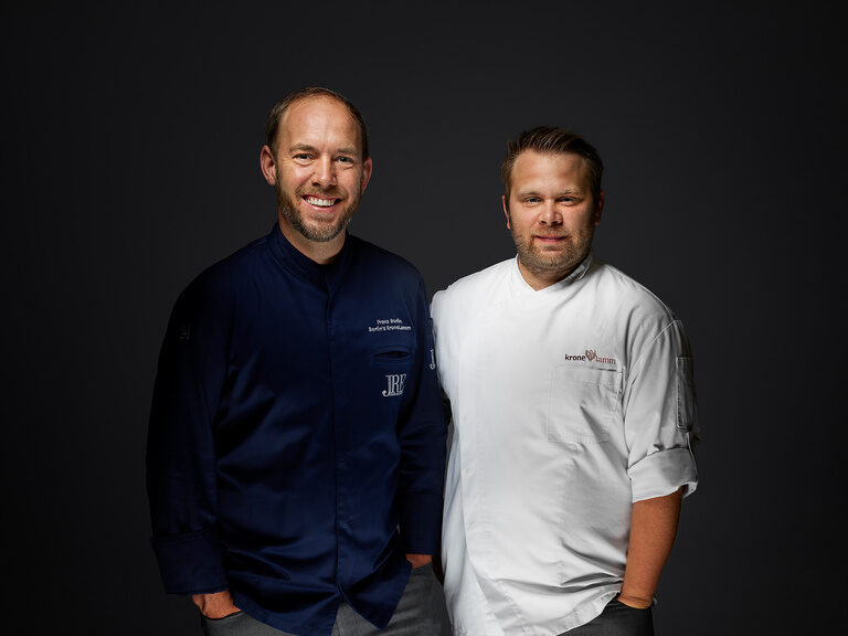 Franz Berlin stands next to Ansgar Blatter in a blue chef's jacket, smiling against a dark background.