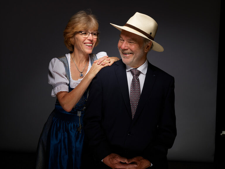 Die Seniorchefs Gudrun und Rolf Berlin setzten, mit dem Kauf des ehemaligen Gasthauses Krone, 1994 den Grundstein für das heutige Hotel Kronelamm im Schwarzwald. Gudrun Berlin im Dirndl und Rolf Berlin im Anzug und mit Panamahut lächeln sich vor einem dunklen Hintergrund an.