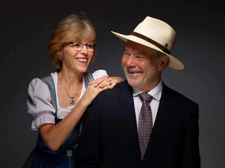 Gudrun Berlin im Dirndl und Rolf Berlin im Anzug und mit Panamahut lächeln sich vor einem dunklen Hintergrund an.