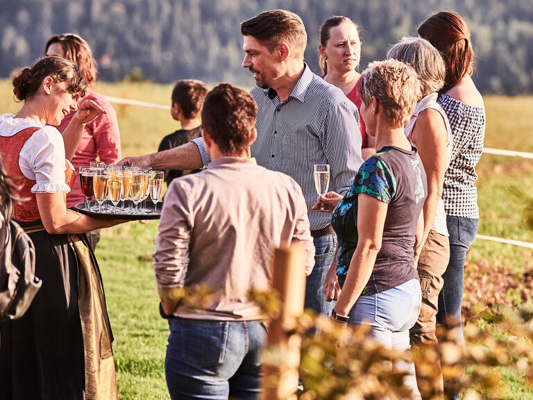 A young service employee holds a tray with champagne glasses in her hand and distributes them to the guests standing around.