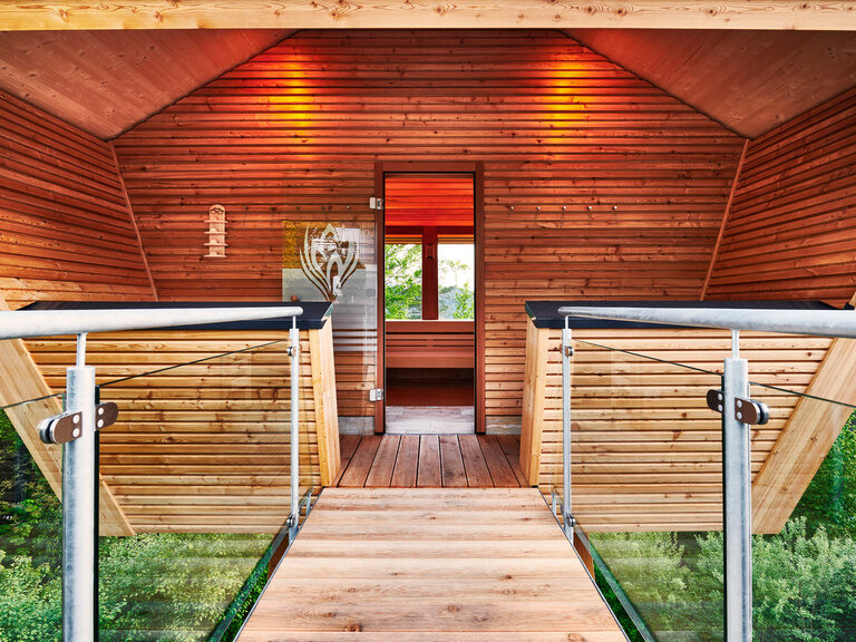 In der Hochsitzsauna entspannen Gäste ungestört und genießen, fernab vom Alltag, ganzheitliche Gelassenheit. Ein hölzener Steg führt zum Eingang einer Sauna, die einem kleinen Haus gleicht.