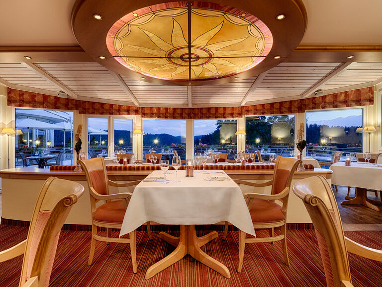 Das Hotelrestaurant Kronelamm bietet das perfekte Ambiente für besondere Anlässe und romantische Stunden zu zweit. Ein gedeckter Tisch mit zwei Stühlen in einer Stube, mit großen Fenstern, durch die die Dämmerung zu sehen ist.