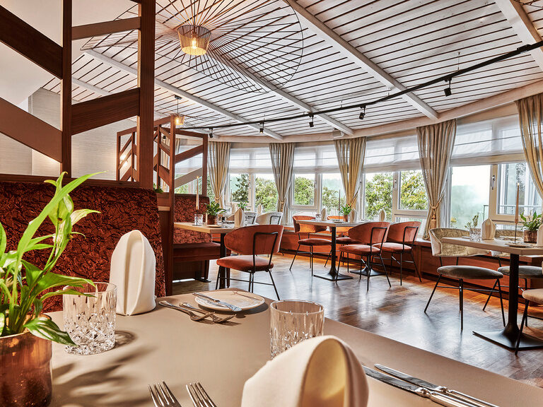 Ein elegant gedeckter Tisch im Restaurant des Hotel Kronelamm, mit Blick auf die großen Fenster und die gemütlichen Sitzbereiche. Die Einrichtung ist modern mit warmen Farbtönen, Pflanzen und viel Tageslicht.