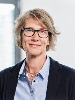 Sympathisch lächelnde Frau mit kurzen, hellen Haaren und Brille. Sie trägt eine hellblaue Bluse und einen dunklen Blazer.