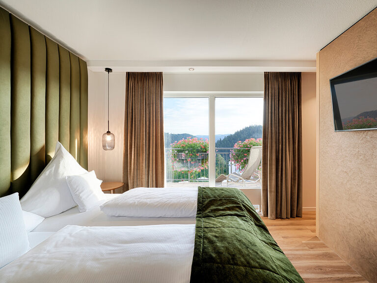 Hotelzimmer der Kategorie Teinachtal im Haupthaus Hotel Kronelamm. Gemütliches Bett mit grüner Tagesdecke vor einem großen Panoramafenster.