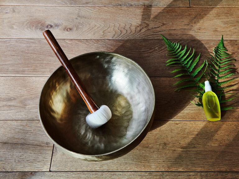 Eine Klangschale mit Schlägel steht auf einem Holzboden. Daneben liegt ein Naturöl auf einem Blatt.