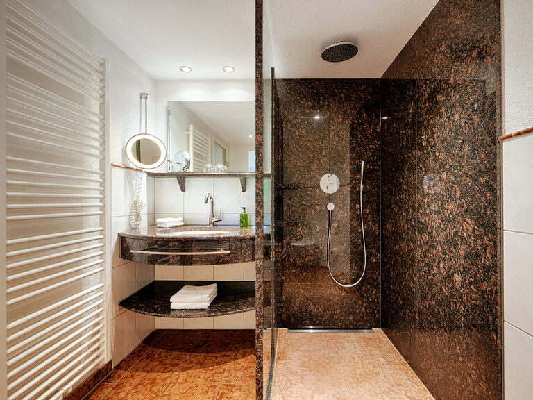 Die beiden Hotelzimmer dieser Kategorie unterscheiden sich in ihrer Einrichtung. Das Badezimmer dieses Zimmers besticht durch eine ebenerdige Dusche. Ein dunkel gehaltenes Badezimmer mit begehbarer Dusche, großem Waschbecken, Spiegeln und Waschutensilien.