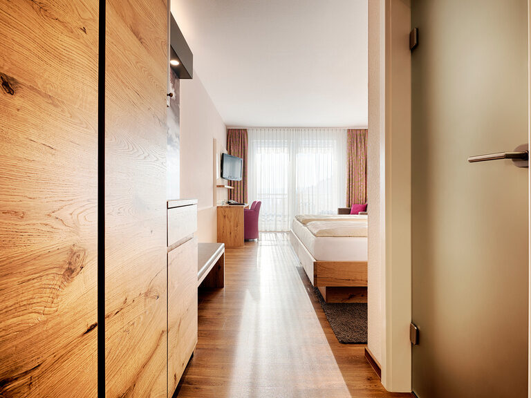 Alle Komfort-Doppelzimmer der Kategorie Poolblick sind modern und komfortabel, ohne Teppichboden eingerichtet. Ein Zimmerflur mit Boden in Holzdesign und Schränken, einer Türe zum Badezimmer und Blick in den Wohnbereich.