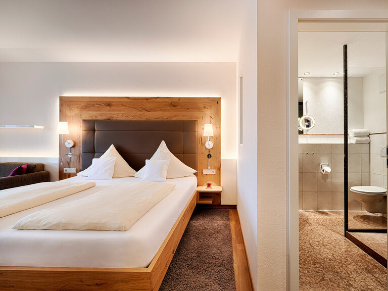 In den Komfort-Doppelzimmern Poolblick im Haupthaus Hotel Kronelamm genießen Gäste einen Blick in den Schwarzwald und über den Außenpool. Ein gemütlicher Schlafbereich mit Holzelementen, der durch eine Wand zum Badezimmer getrennt ist.