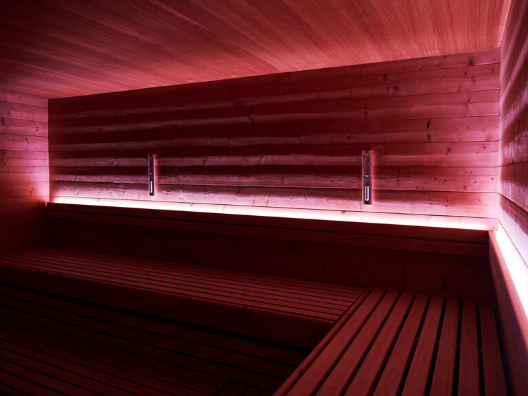 Die Kräutersauna des Hotels in Zavelstein ist aufgrund des milden Raumklimas besonders bei Sauna-Neulingen beliebt. Sitzbänke einer Sauna werden rosa beleuchtet.