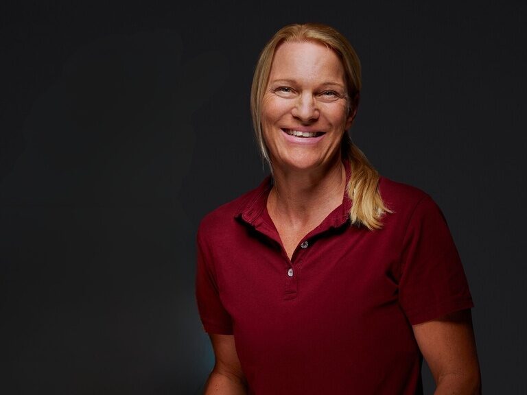 Kristina Möhrer ist Fitnesstrainerin und steht Gästen motivierend zur Seite. Sporttrainerin Kristina Möhrer lächelt bei einem Portraitfoto vor dunklem Hintergrund.