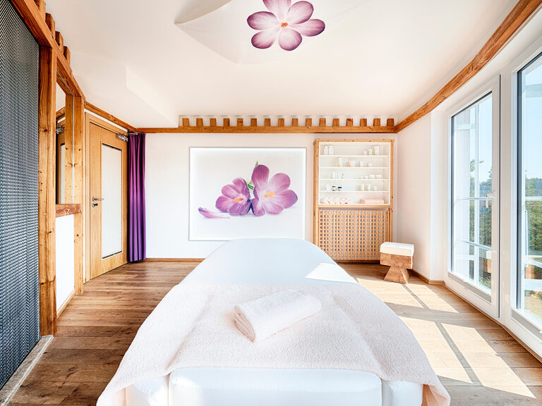 Der Blick von einer weißen Massageliege auf ein Bild mit einer Blüte in einem geräumigen, lichtdurchfluteten Raum.