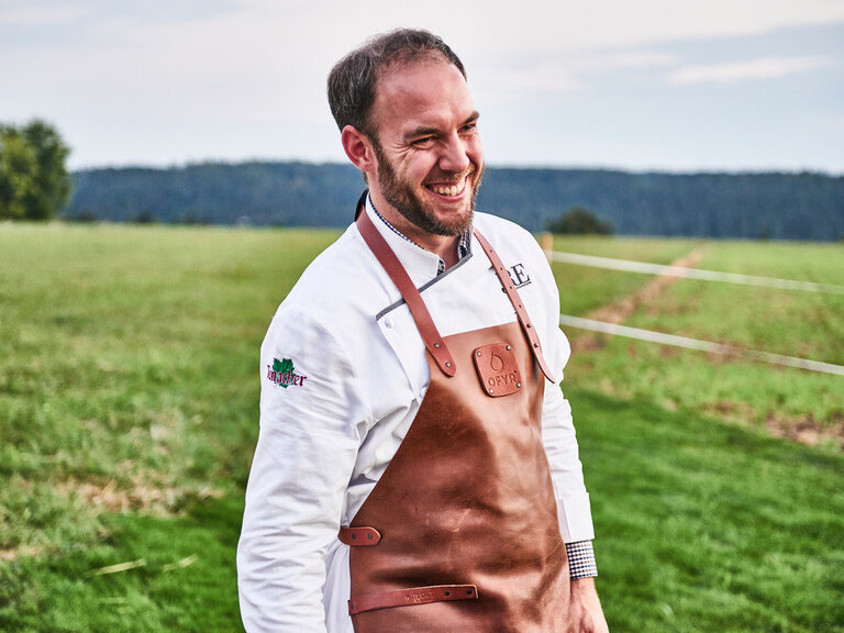Franz Berlin steht nicht nur in seinem Gourmetrestaurant in der Küche, sondern oftmals auch bei Events im Wanderheim oder auf dem Theurerhof. Franz Berlin trägt eine weiße Kochjacke, eine braune Lederschürze und steht lächelnd auf einer grünen Wiese