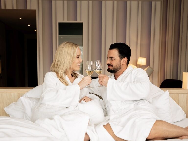Im Hotel Kronelamm wird Ihr Besuch zu einem unvergesslichen Erlebnis. Ein lächelndes Paar in Bademänteln stoßt in einem Bett im Wellnesshotel Kronelamm im Schwarzwald mit zwei Gläsern Sekt an.