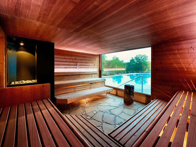 Die etwa 70°C warme Panorama-Softsauna mit wunderbarem Ausblick auf Außenbereich und Outdoorpool des Wellnesshotels. Rotbeleuchtete Sauna mit Sitzgelegenheiten aus Holz und großem Panoramafenster mit Blick auf den Außenpool.