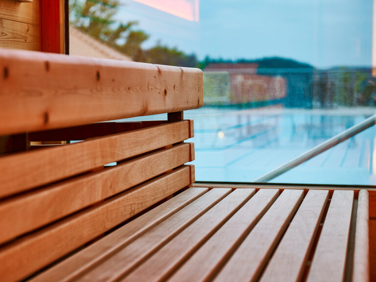 Das Wellnesshotel Kronelamm im Schwarzwald ist aufgrund des großen, erholsamen Wellnessbereichs sehr beliebt. Sitzbänke in einer Sauna, die durch ein Fenster vom Pool getrennt sind.