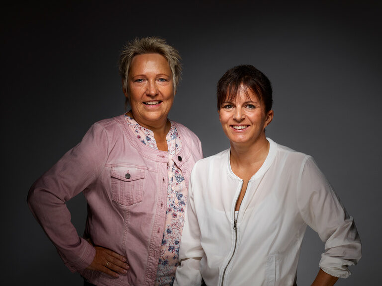 Das Verwaltungs-Duo Petra Volle und Anita Nothacker kümmert sich um alle Belange rund um Mitarbeiter:innen und Buchhaltung. Petra Volle und Anita Nothacker lächelnd vor einem dunklen Hintergrund.