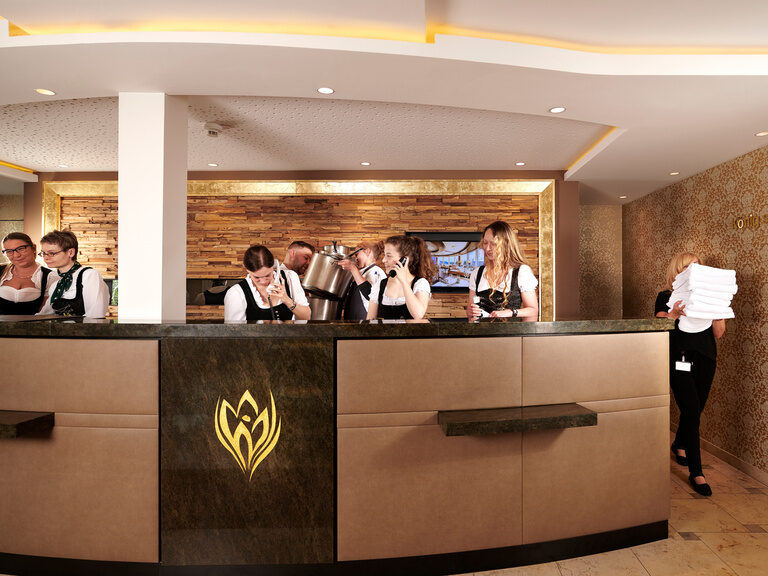 Die Hotelrezeption des Hotels Kronelamm im Schwarzwald steht allen Gästen bei Fragen, Wünschen und Anregungen jederzeit zur Verfügung. An einer Hotelrezeption befinden sich viele Mitarbeiter:innen, die telefonieren, arbeiten, lachen und Spaß haben.