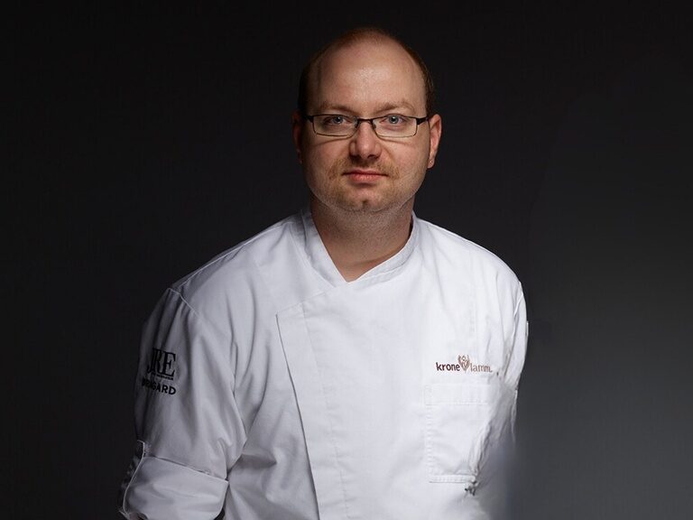 Seit 2008 gehört Robby zum Team des Hotel Kronelamm und verwöhnt die Gäste im Wanderheim mit liebevoll zubereitetem Essen und leidenschaftlichem Engagement.. Der Wanderheim Küchenchef Robby Mittag posiert vor dunklem Hintergrund für ein Portraitfoto.