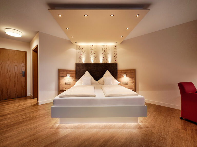 A white, illuminated bed and a red armchair in a hotel room with wooden floors and wooden doors.