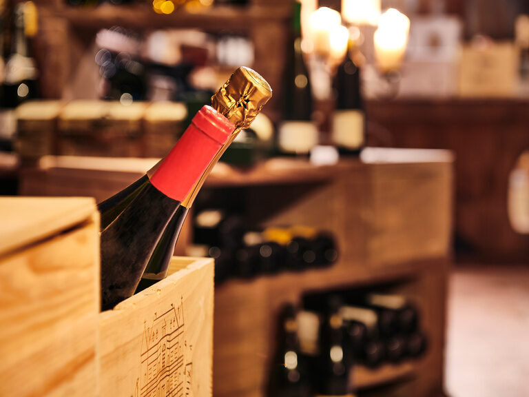 Zwei Sommeliers, Roland Berlin und Michael Kolb, garantieren eine exzellente Weinauswahl und runden damit jedes Gericht des Küchenteams ab. Nahaufnahme einer Wein- und einer Sektflasche die in einer Holzkiste stehen