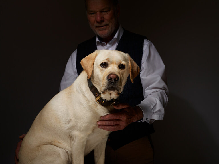 Rolf Berlin, Seniorchef des Wellnesshotels Kronelamm in Zavelstein, ist fast immer mit seinem Hund Prinz anzutreffen. Seniorchef, Rolf Berlin, und sein weißer Hotelhund Prinz vor einem dunklen Hintergrund.