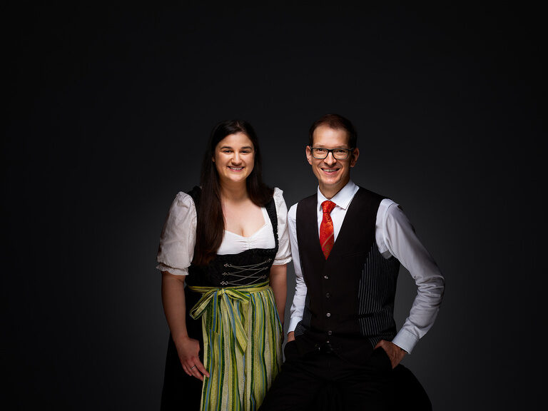 Stellvertretende Empfangsleiterin Simona Festa und Empfangsleiter Oliver Kopp sind jederzeit persönliche Ansprechpartner der Gäste. Simona Festa und Oliver Kopp lächeln in Arbeitskleidung vor einem dunklen Hintergrund.