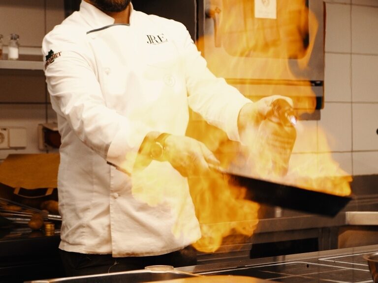 Franz Berlin wears a chef's jacket and holds a pan that throws flames in his hand.