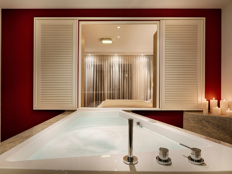 Whirlwanne in der Kronelamm Suite des Wellnesshotels Kronelamm im Schwarzwald! Eine mit Wasser befüllte Whirlwanne, die vor einer Glasfront mit Blick in ein Schlafzimmer steht.
