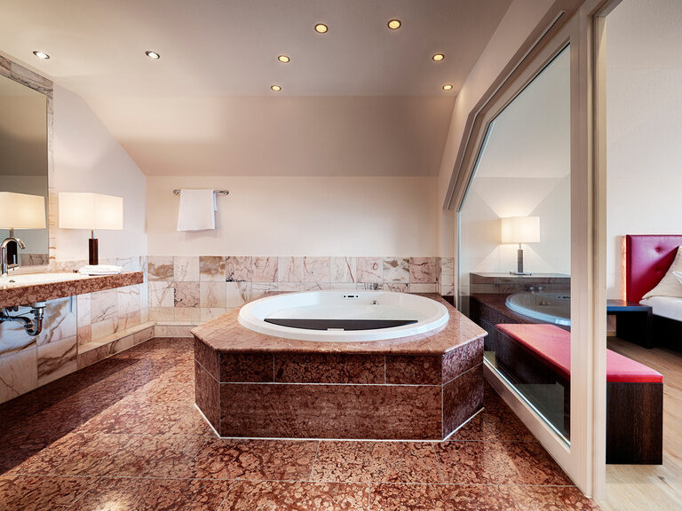 Das Badezimmer der Zavelstein Suite ist mit luxuriösem Portugalmarmor ausgestattet, verfügt über eine große Dusche, Whirlpool, Doppelwaschbecken und eine separate Toilette. Ein großer Whirlpool in einem Badezimmer, welches durch eine Glasfront zum Schlafbereich getrennt ist.