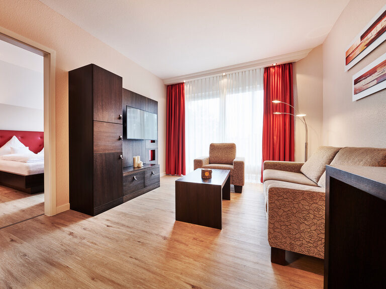 Die Zavelstein Suite befindet sich zur Ostseite im Hotel Kronelamm im Schwarzwald mit Blick auf das historische Zavelsteiner Städtle. Ein Wohnbereich mit großer Couch, dunkler Wohnwand aus Holz und Fernseher mit roten Elementen.