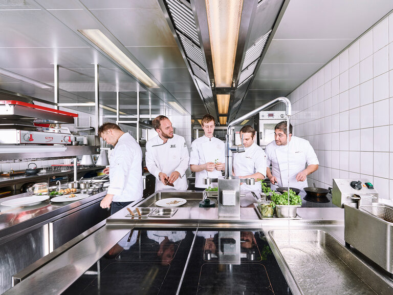 Franz Berlin und sein Küchenteam bereiten kulinarische Highlights, in der Küche des Gourmetrestaurants Berlins Krone, zu. Fünf Köche arbeiten mit Freude in einer geräumigen Restaurantküche.