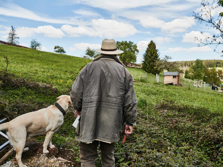 Rolf Berlin and his white Labrador stand on a green meadow under a blue sky and look into the distance.