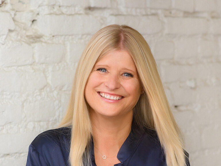 Eine sympathisch lächelnde Frau mit langen blonen Haaren. Sie trägt eine blaue Bluse und steht vor einer weißen Steinwand.