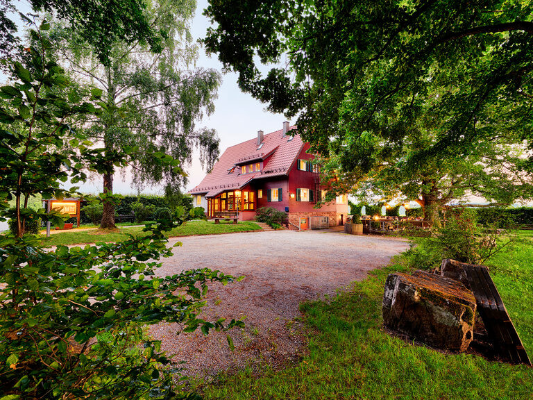Große Biertische stehen in einem Biergarten vor einem roten Haus.