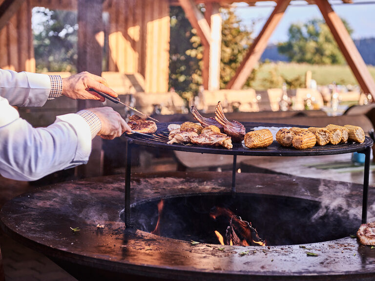 Auf einem großen Grill liegen verschiedene Fleischstücke sowie Maiskolben, die von einem Koch zubereitet werden.