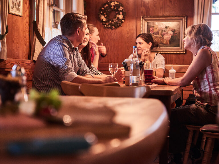 An einem rustikalen Tisch in einer Stube sitzen drei Personen, die sich unterhalten.